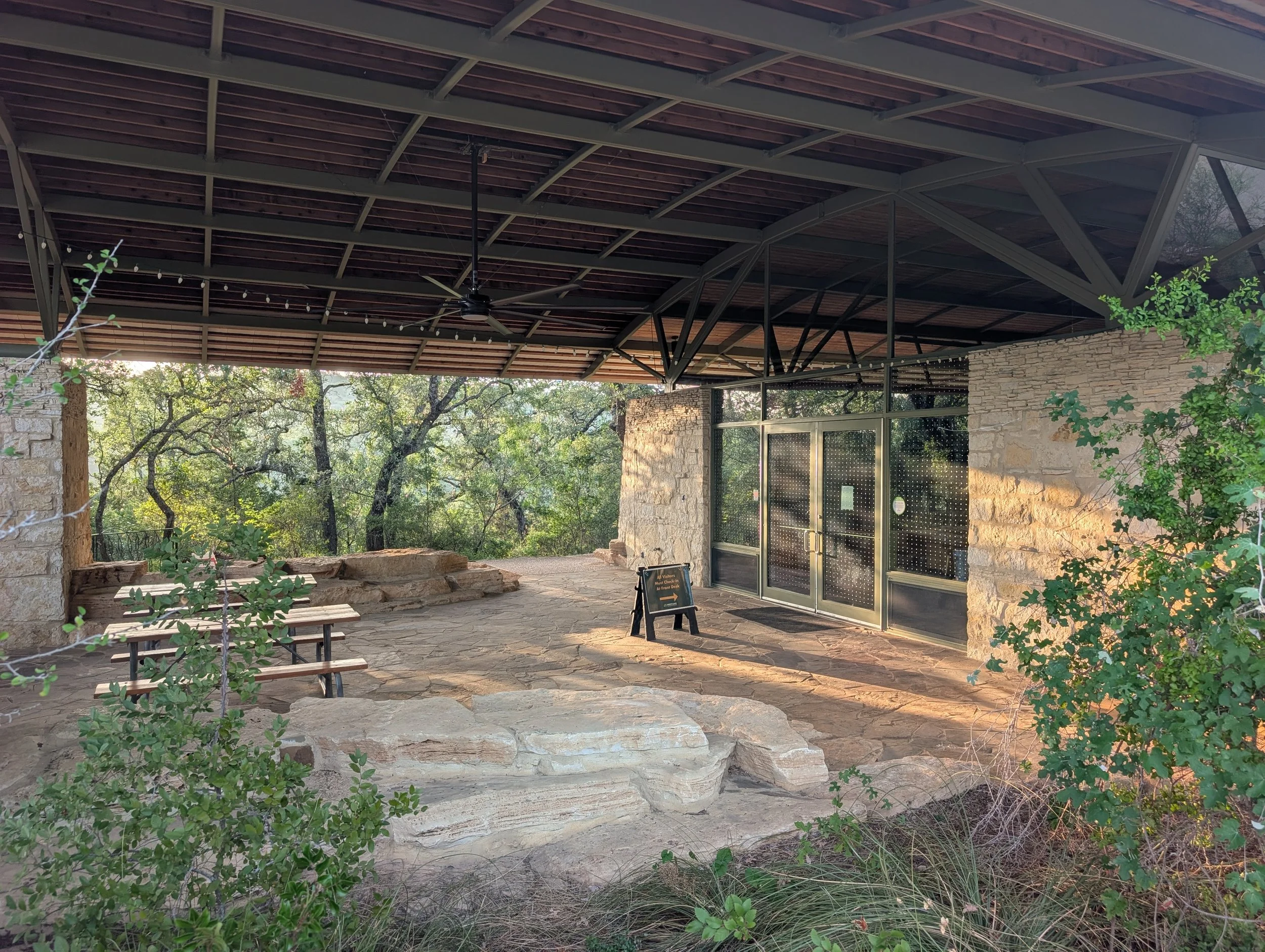 Outdoor patio area at the environmental learning center with stone walls, metal and glass door, wooden benches, plants, and string lights under a metal roof.