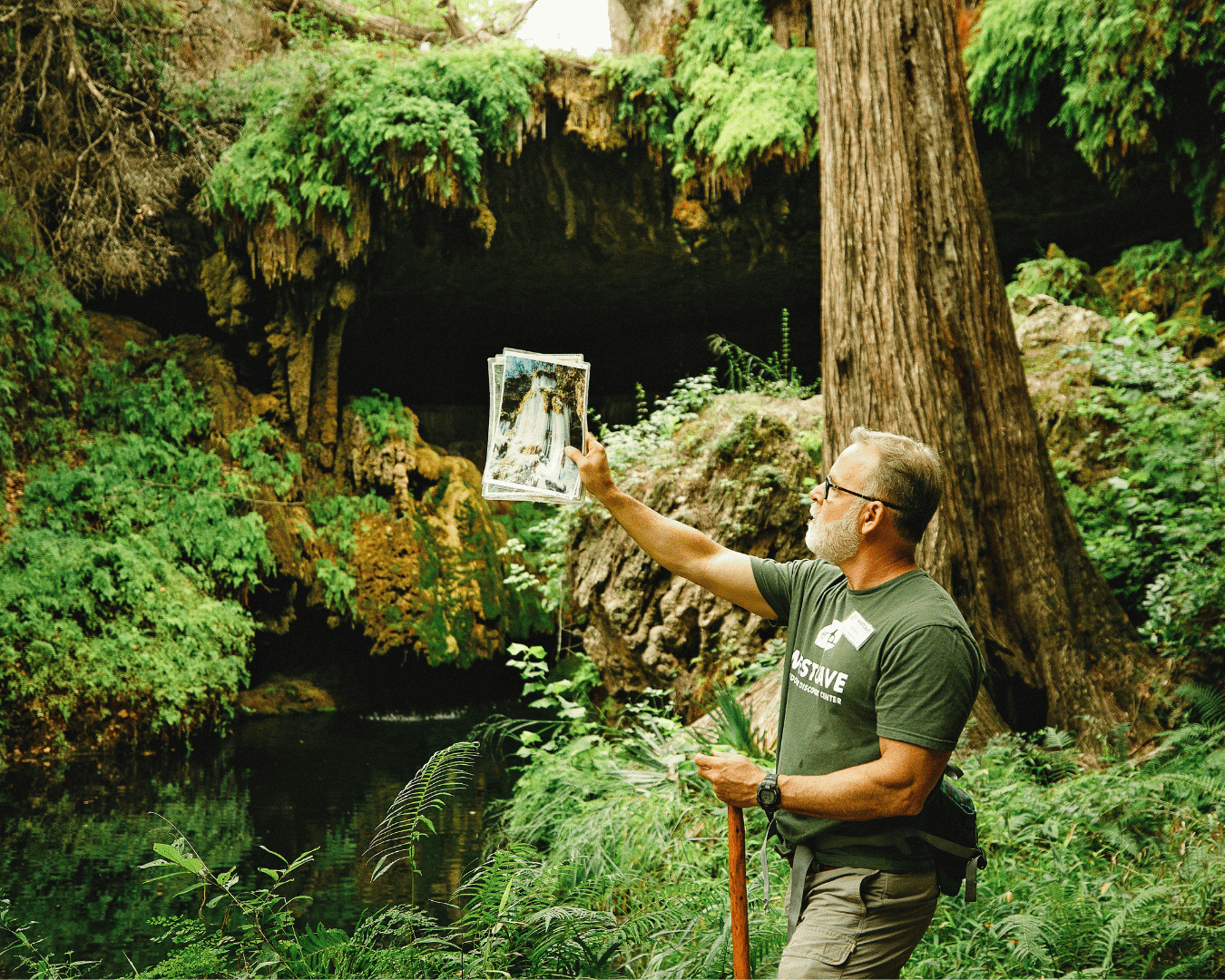 A man with glasses and a beard in a forest holding up a photograph of a waterfall.