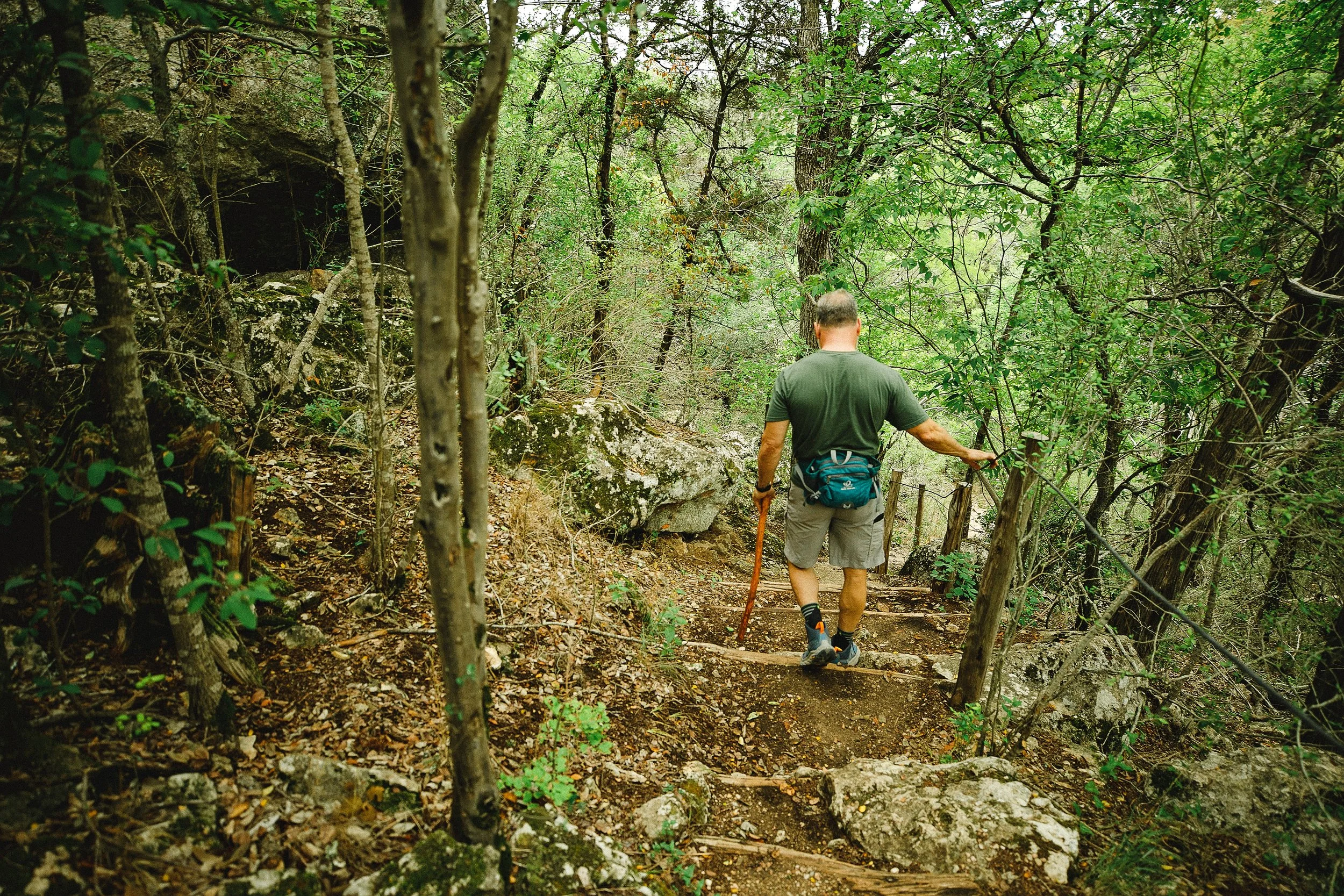 A man hiking through a dense, green forest, wearing a green t-shirt, shorts, and hiking shoes, carrying a backpack, and using a walking stick.