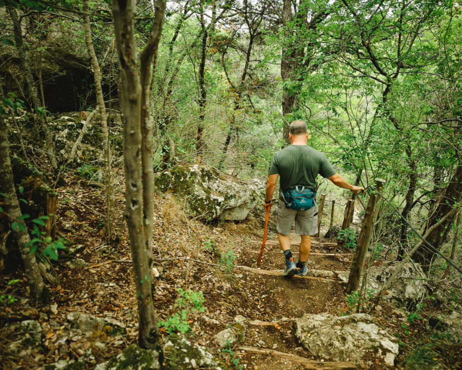 A man in a green t-shirt and gray shorts walking down a forest trail with a walking stick, surrounded by trees and rocks.