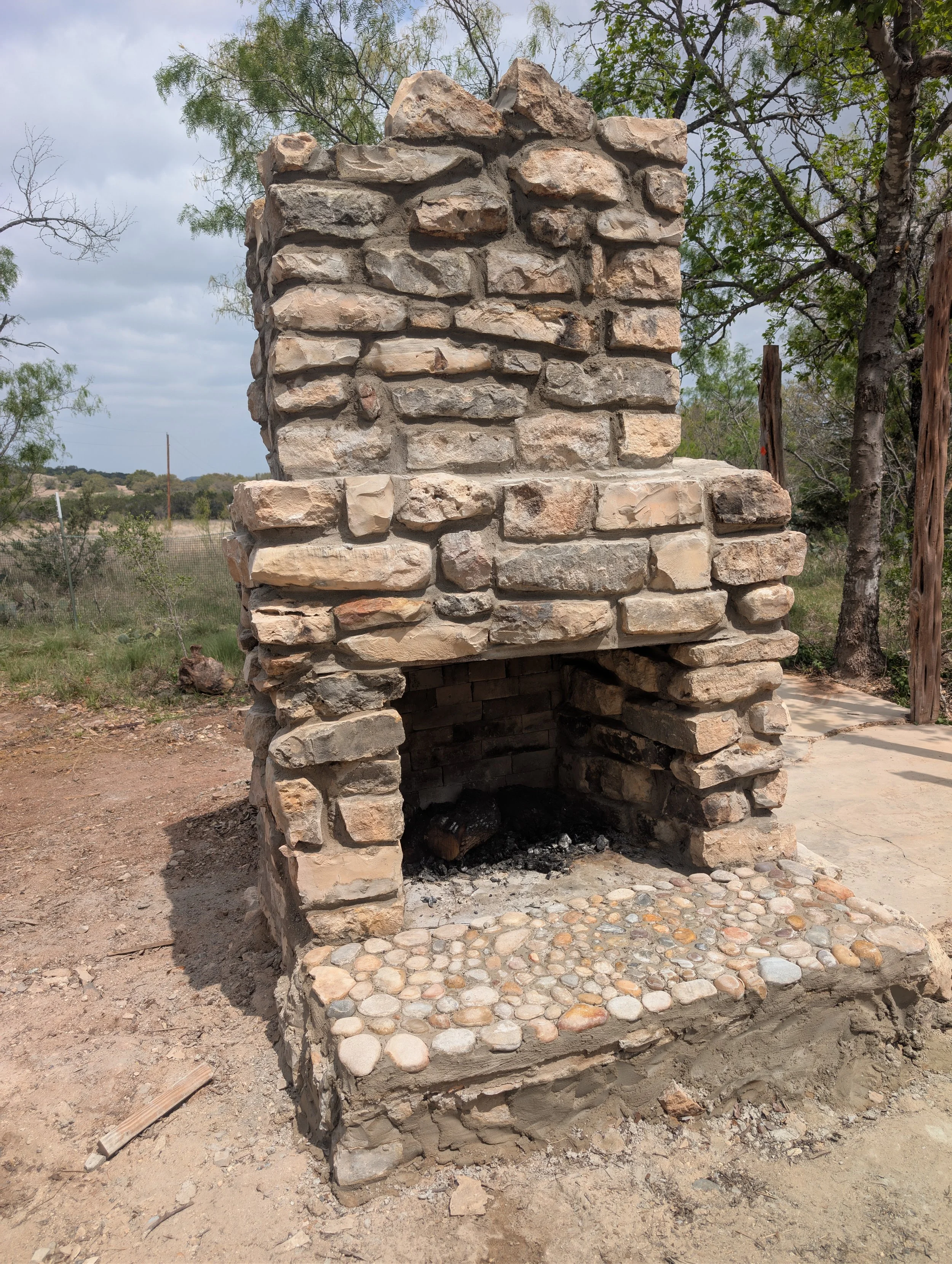 A rustic outdoor fireplace made of stacked irregular stones with a firebox at the bottom and a chimney above. There are trees in the background and a partially cloudy sky.