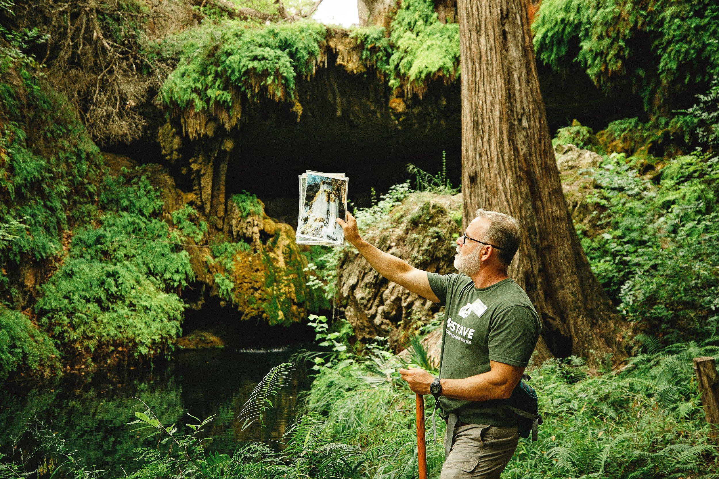 A man with glasses and a beard holding a photo of a waterfall in front of a lush green cave. He is wearing a gray t-shirt and khaki shorts, standing near a body of water in a forest setting.