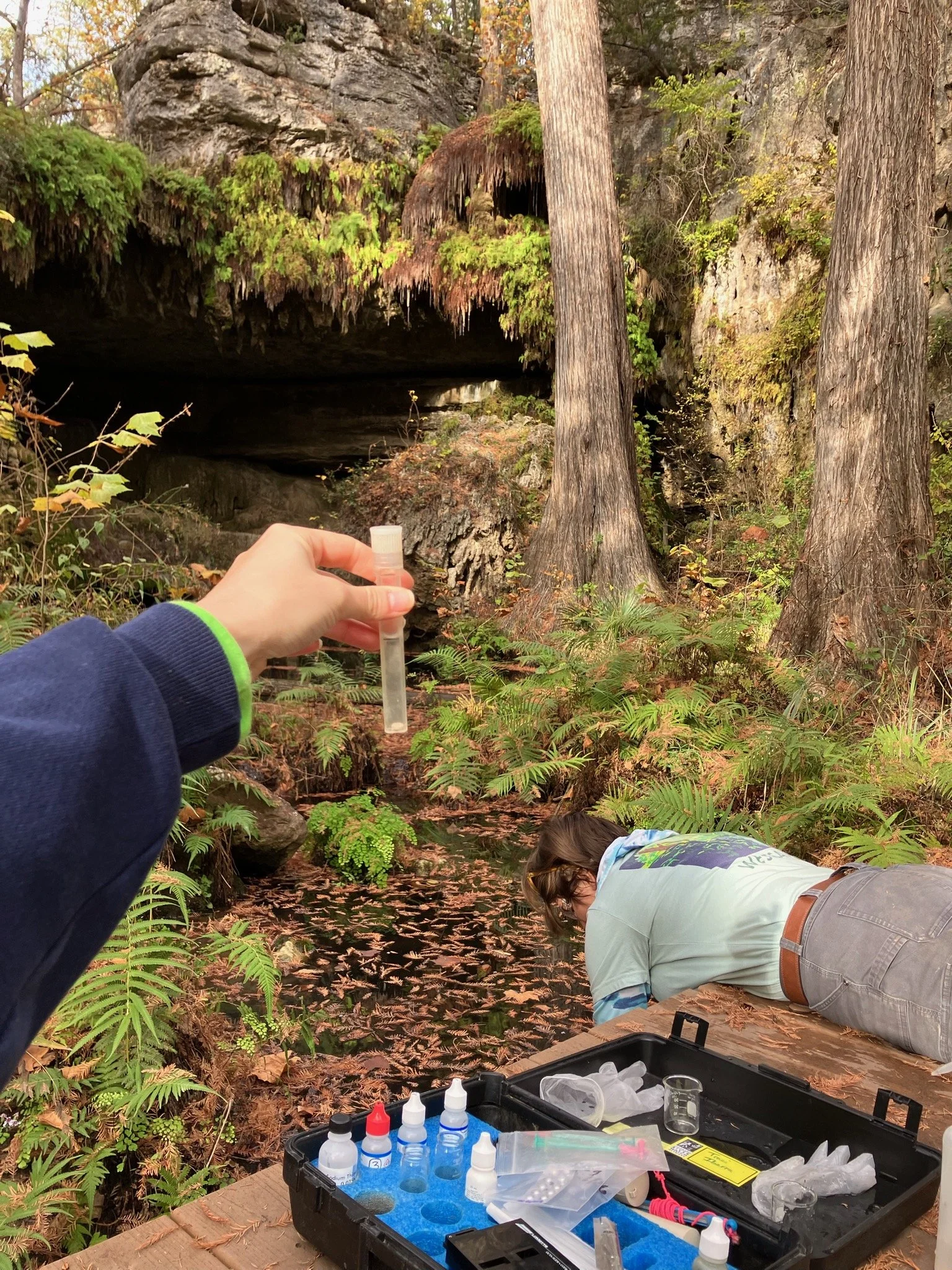 A person in a light gray shirt and khaki pants is crouched down near a small stream in a forest, with a backpack in front. Another person, only partially visible, is holding a small clear tube or vial, likely collecting a sample. A black case with various bottles, containers, and tools for scientific work is opened on a wooden surface nearby, with a background of trees, rocks, and leafy ground.