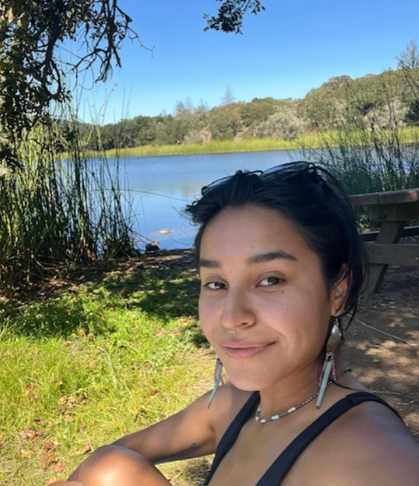 A woman with black hair, a lake and greenery in the background.