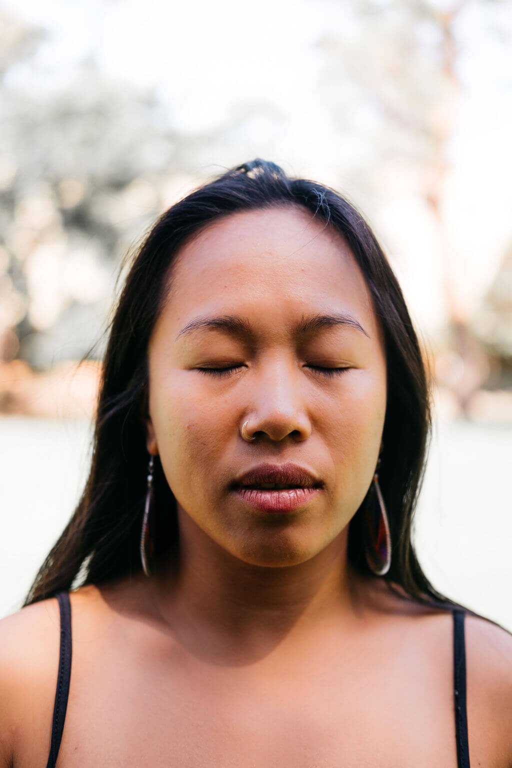 Close-up of a young woman with closed eyes, wearing a nose ring and earrings, outdoors with blurred trees in the background. She is practicing Vedic Meditation.