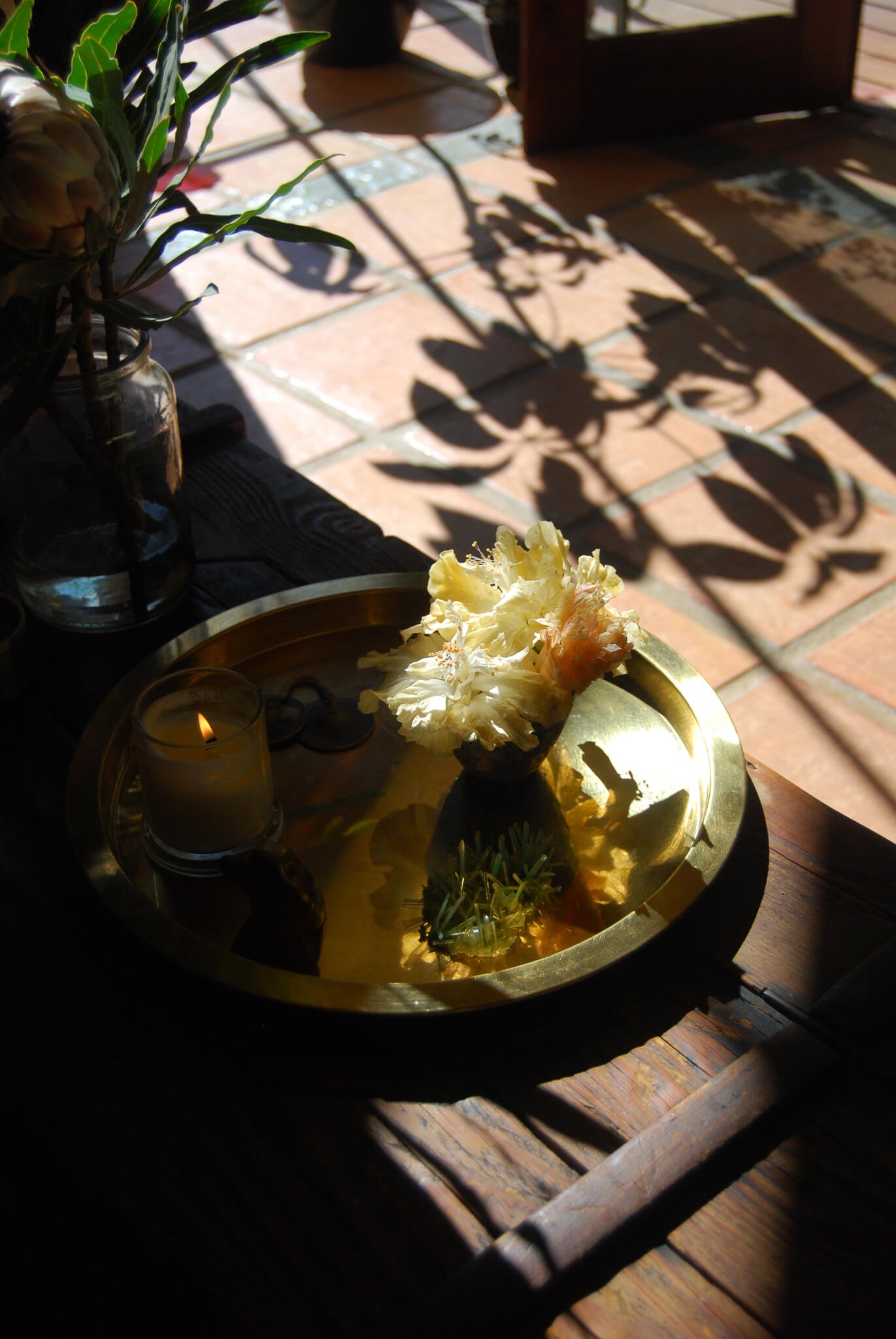 A brass tray with a white candle, a small plant, and a flower arrangement sitting on a wooden table next to a window with sunlight and shadows on the tiled floor.