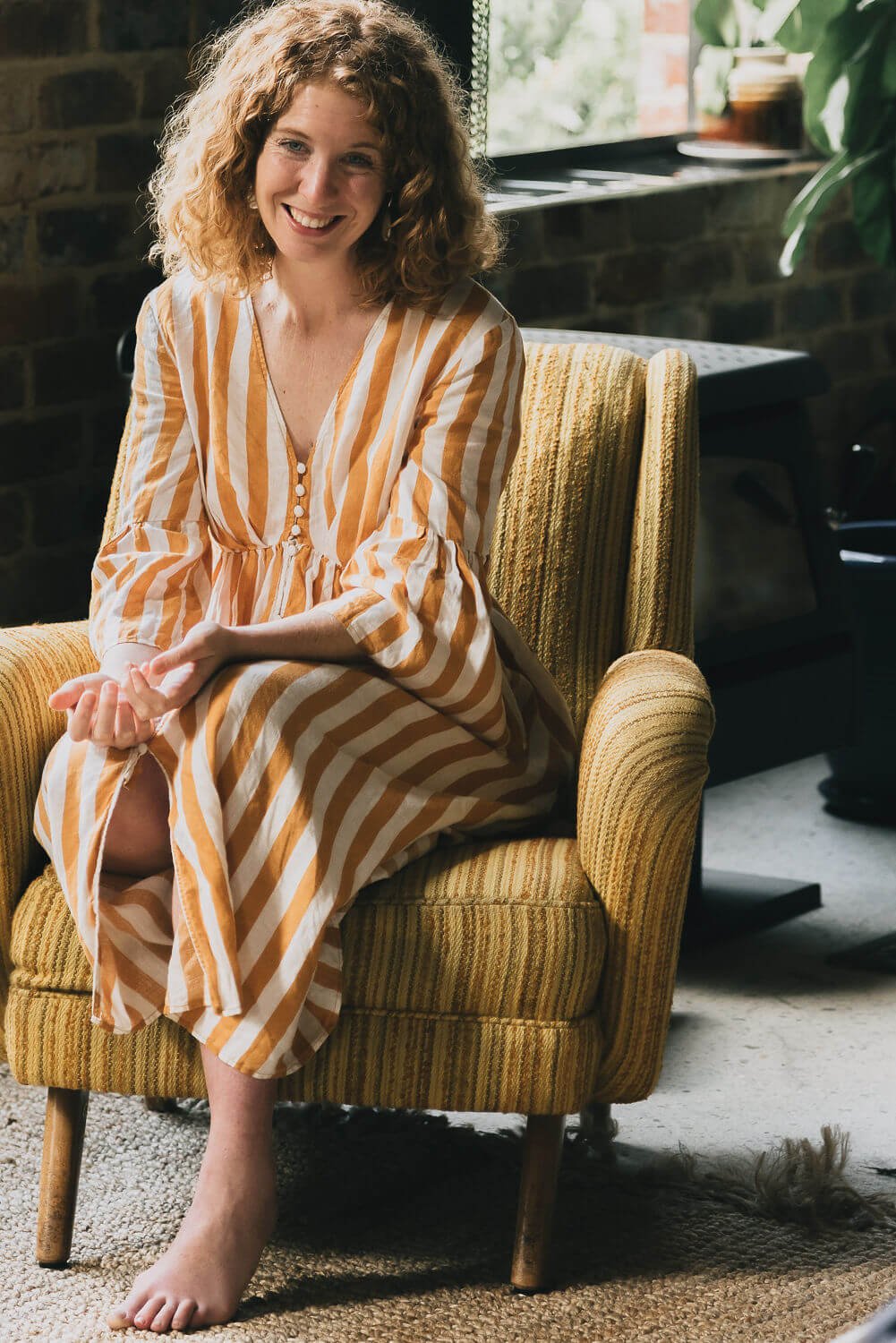 Woman with curly hair smiling, sitting on a yellow striped armchair in a cozy room with a brick wall and large window, wearing a yellow and white striped dress.