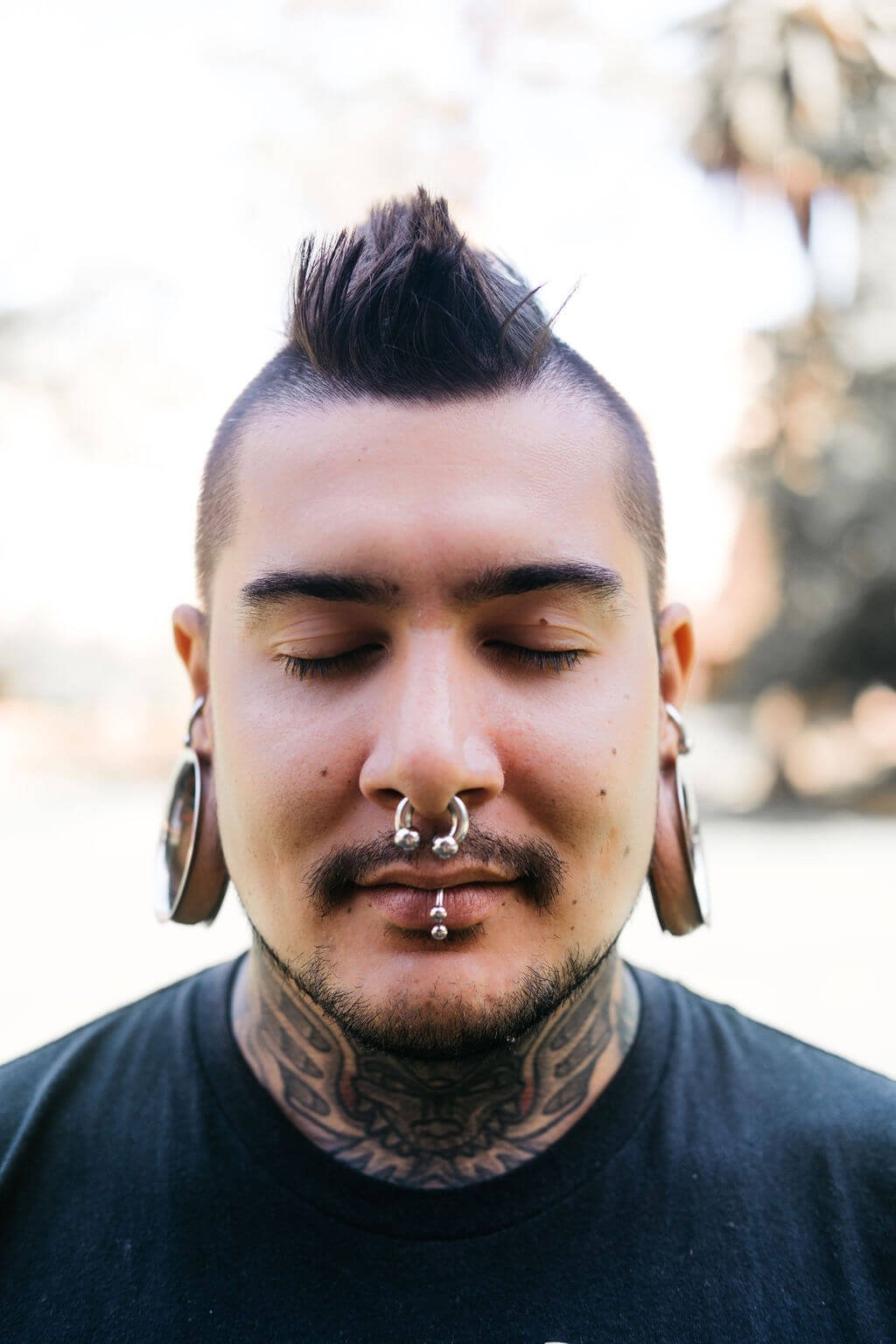 Close-up of a young man with closed eyes, dark hair styled in a mohawk, multiple facial piercings including a nose ring and lip piercings, large earrings, and tattoos on his neck.. He is practicing Vedic Meditation.