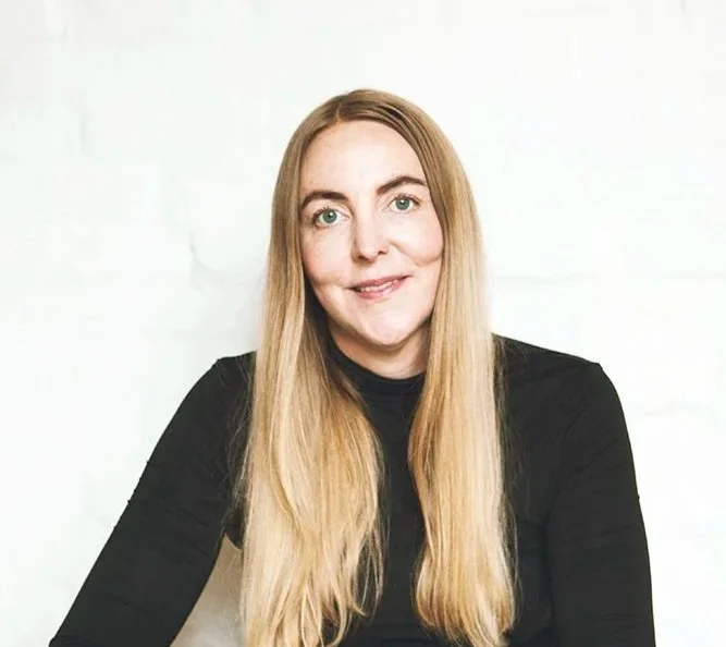 En kvinde med langt blond hår og grønne øjne, smilende, iført sort tøj, foran en hvid væg.