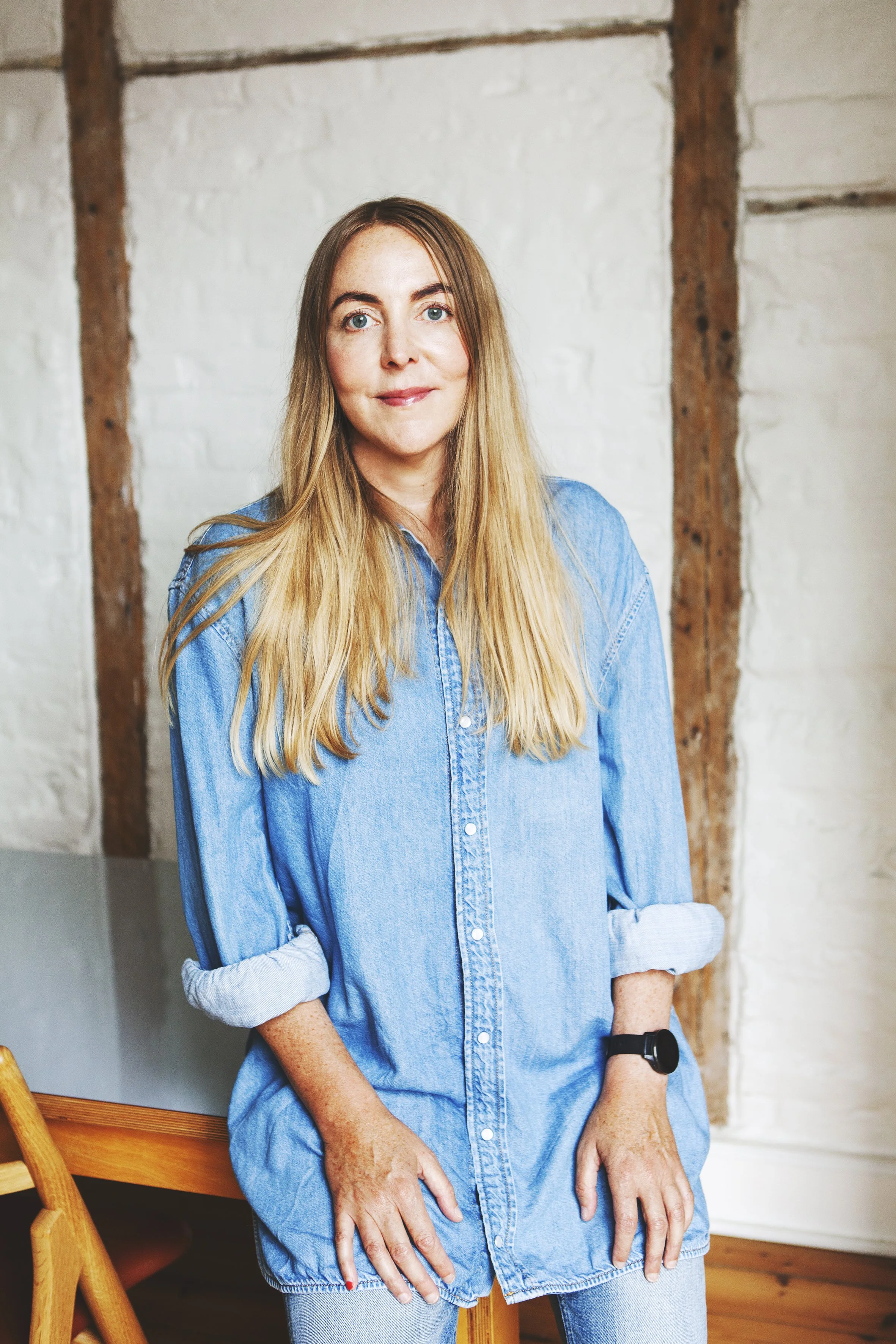 En kvinde med langt blond hår iført en blå denim skjorte, står indendørs foran en hvid væg med træbjælker.