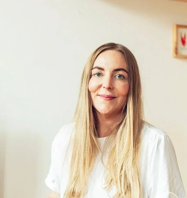 Et portrætbillede af en kvinde med langt blondt hår, iført hvidt tøj, siddende i et indendørsmiljø.