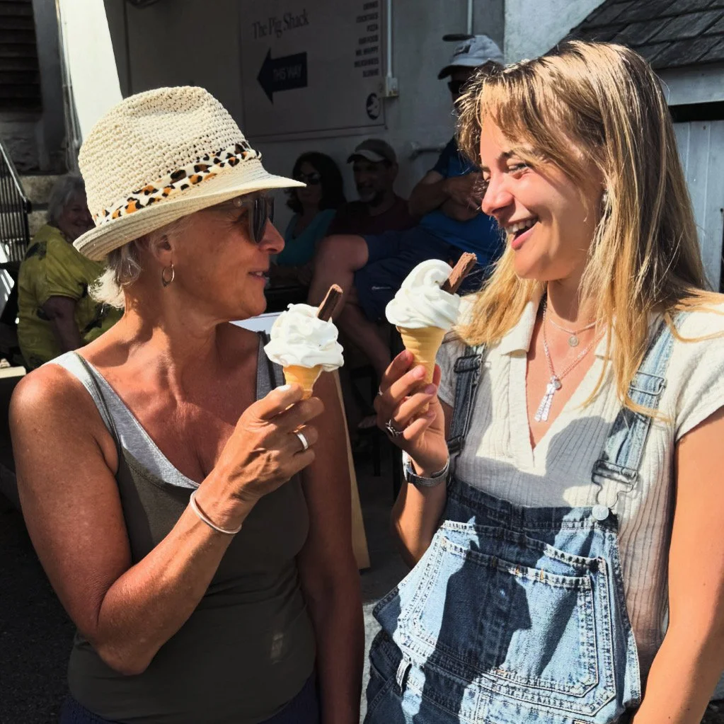 Two women smiling at each other while holding ice cream cones with whipped cream and chocolate sticks. They are outdoors, and there are people sitting in the background.