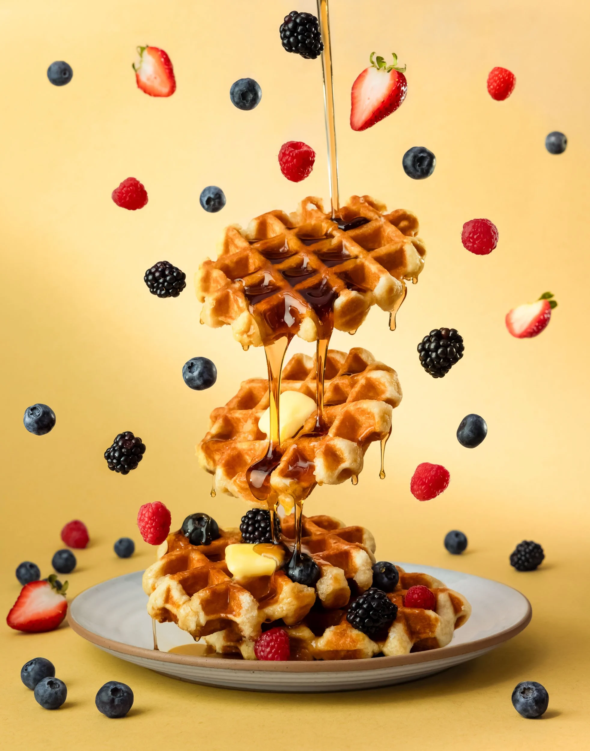 Commercial food levitation photography, creative waffle stack with pouring maple syrup, high-speed composite food styling, professional breakfast advertising imagery, flying berries and butter culinary art for CPG branding.