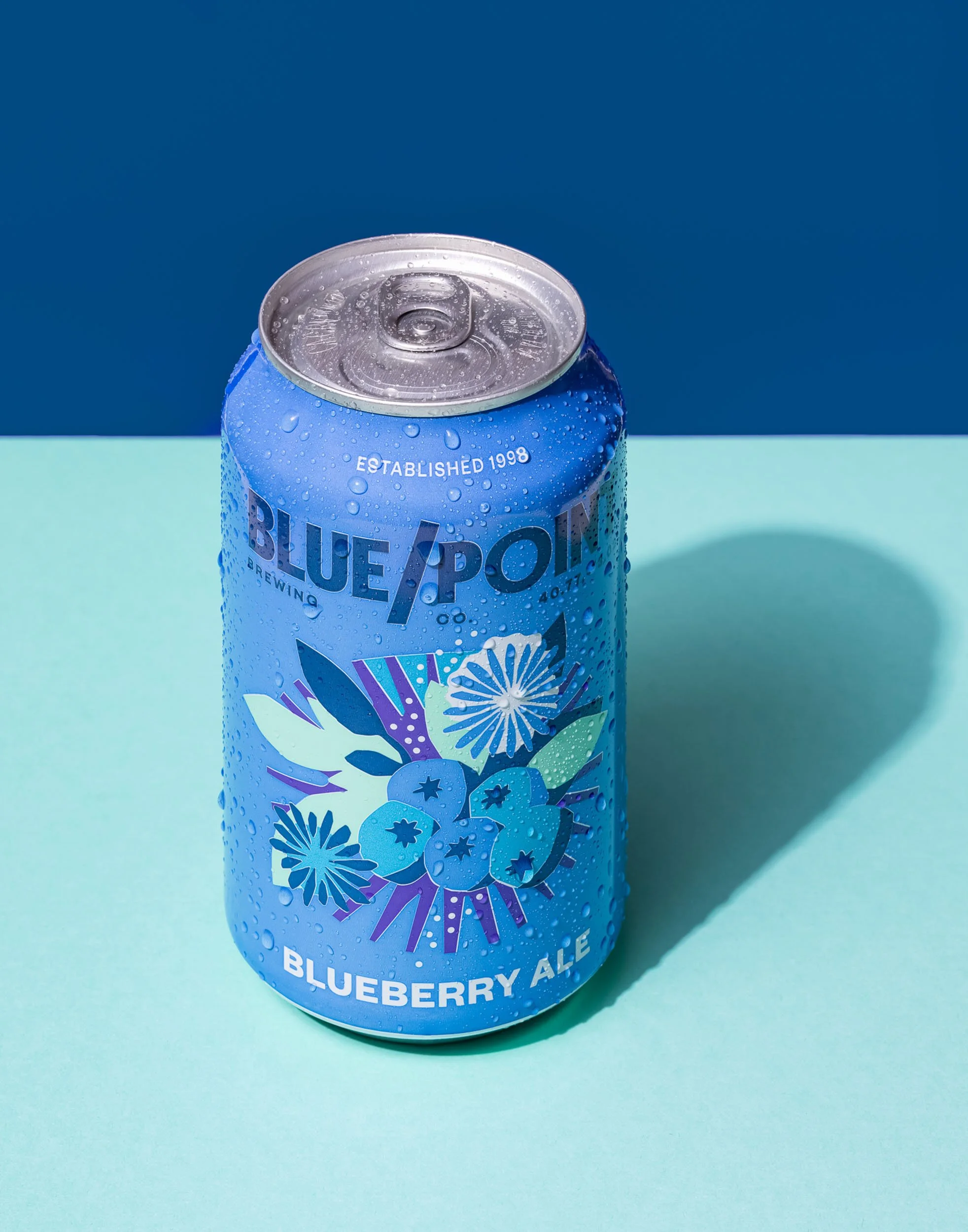 Blue Point Blueberry Ale product photography, teal and dark blue monochromatic beverage branding, seasonal fruit beer commercial shot, refreshing blueberry ale can imagery, modern craft beer e-commerce photography.