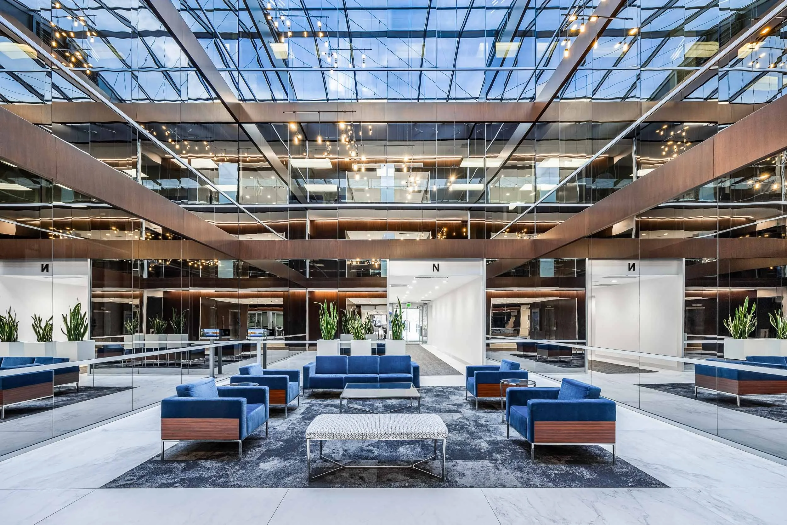Modern commercial atrium interior photography, luxury corporate lobby with glass walls and reflective surfaces, professional architectural lighting for expansive glass structures, high-end office lounge with skylight and blue accent seating, complex 
