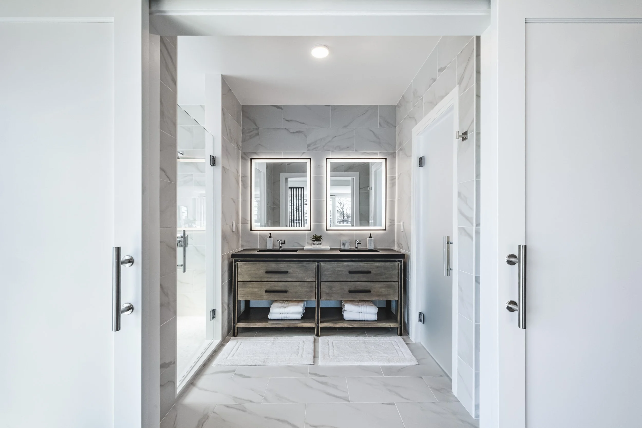 Luxury primary bathroom architectural photography, professional shot of modern double vanity with backlit mirrors, symmetrical interior design with marble tiling and black hardware, upscale hospitality bathroom photography, minimalist luxury interior
