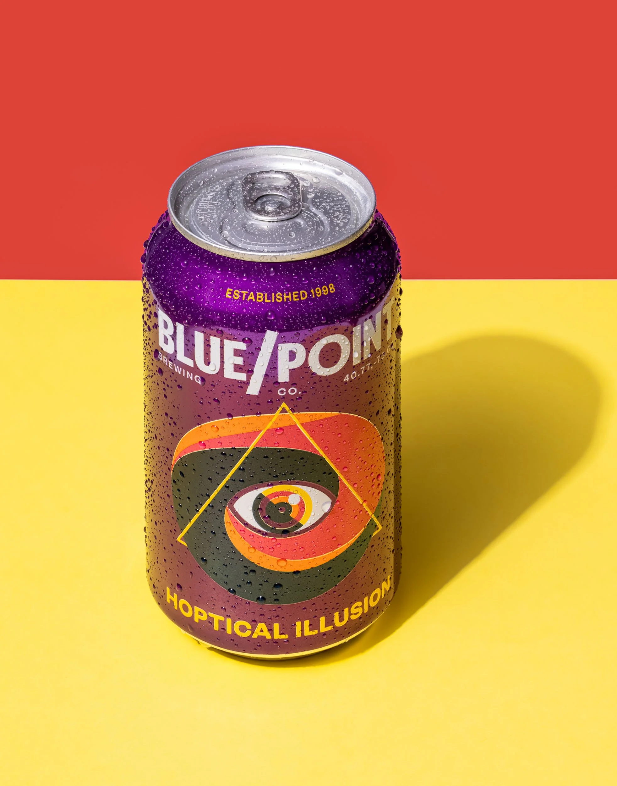 Blue Point Hoptical Illusion IPA product photography, yellow and red vibrant beverage styling, craft beer IPA branding imagery, psychedelic label design commercial shot, professional beer can condensation detail.