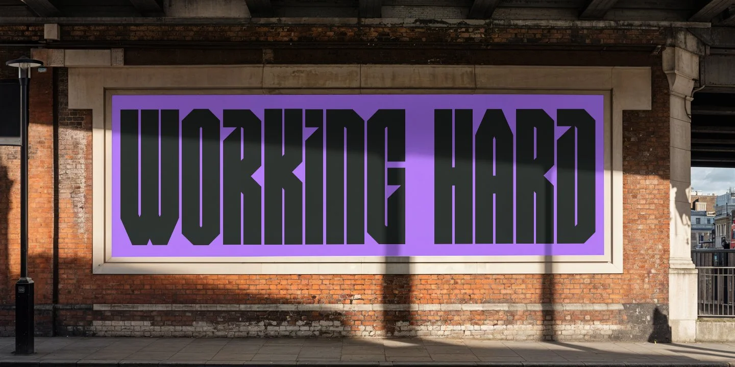 A large electronic billboard displays the words 'WORRING HARD' in bold black letters on a purple background, mounted on a brick wall in an outdoor urban setting.