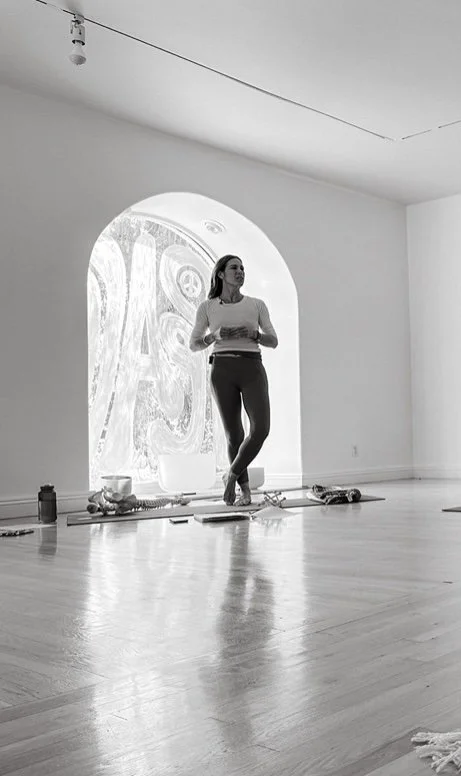 A woman standing in an art gallery or studio space near a large window, holding a cup, surrounded by art supplies, with a large abstract artwork on the window and a high ceiling with a light fixture.