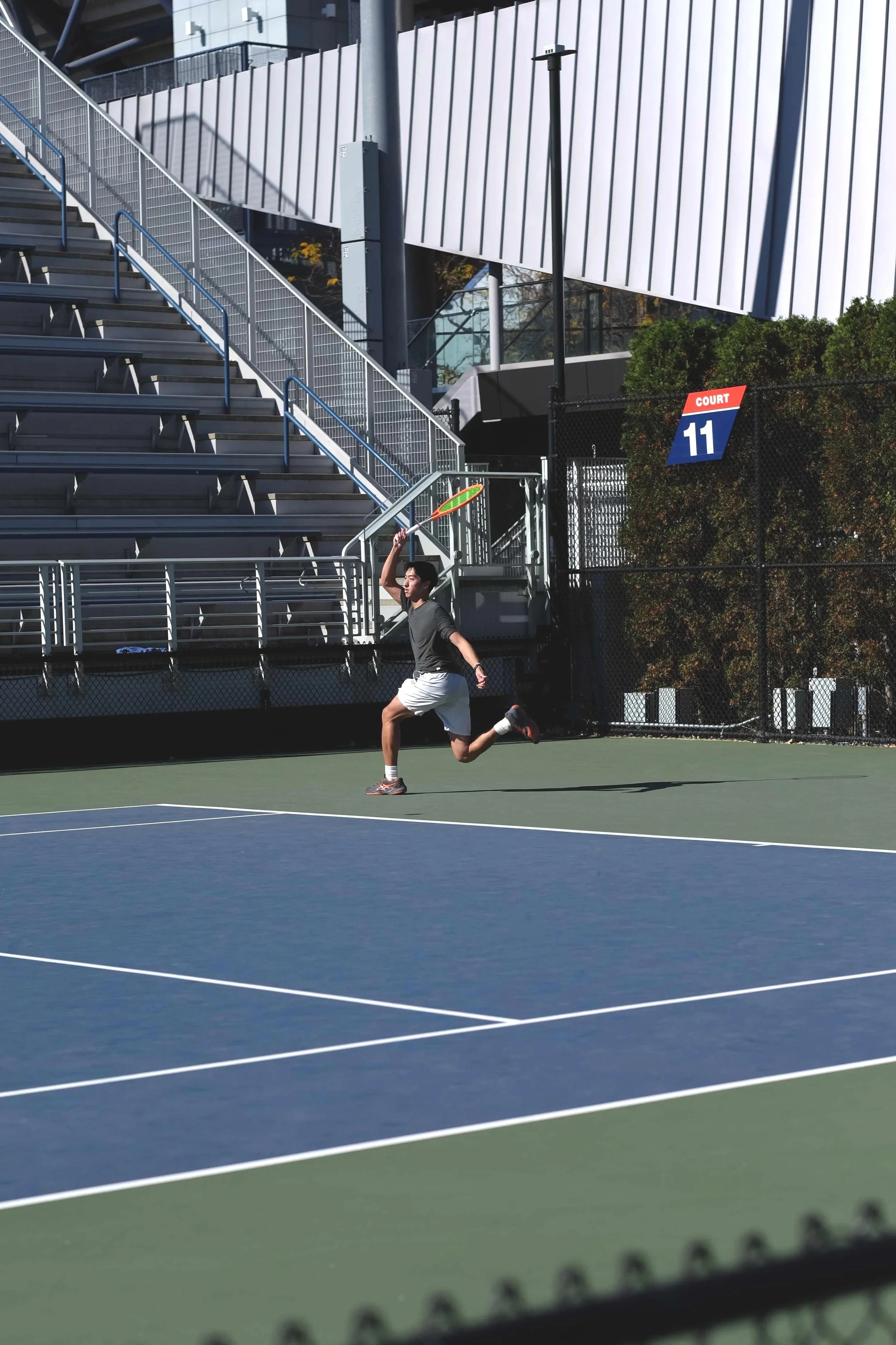 A person playing tennis on an outdoor court with a tennis racket in hand, preparing to hit the ball. The court is surrounded by empty bleachers and a sign that reads 'Court 11'.