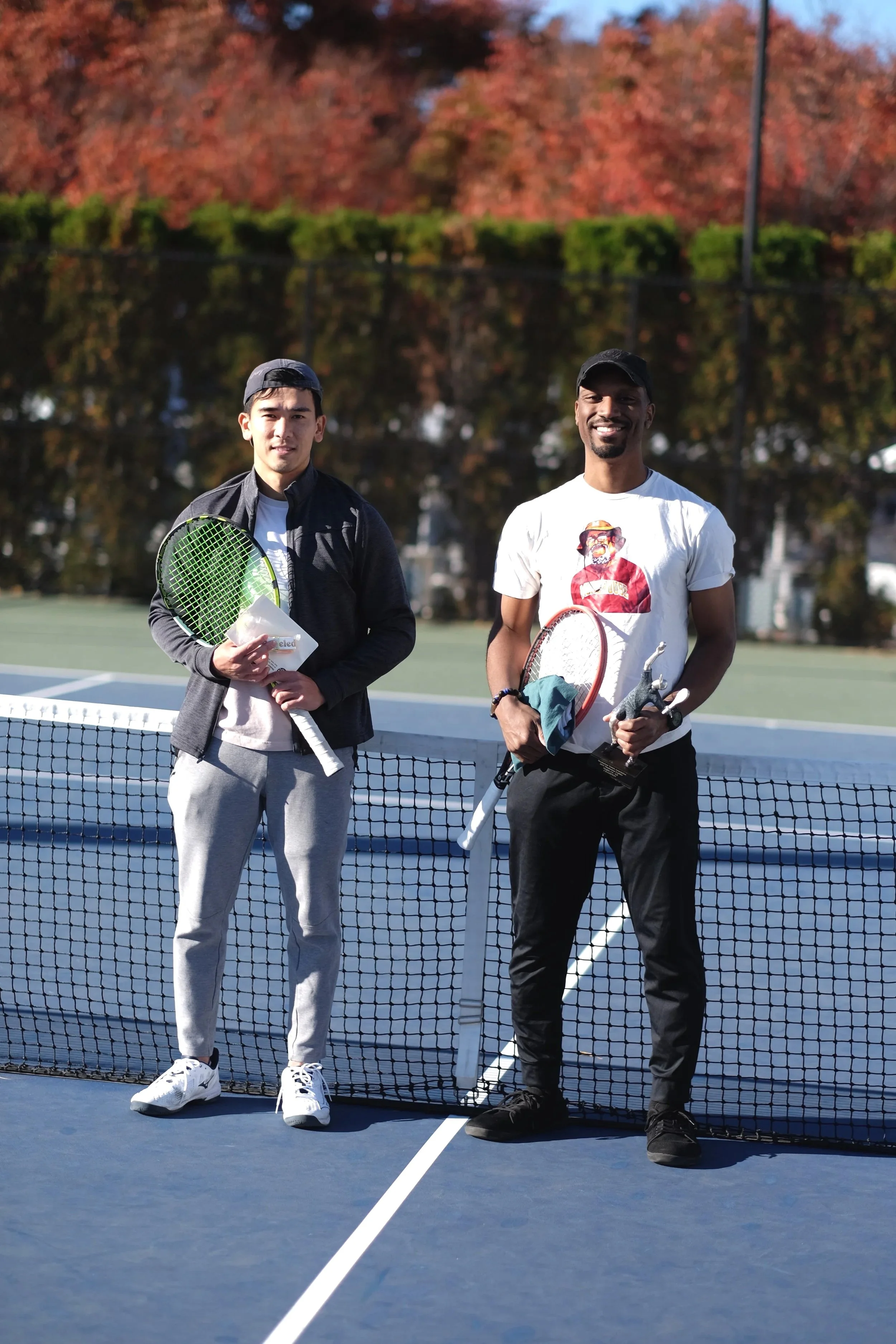 Two men standing on a tennis court holding tennis rackets, with trees in the background and a net in front.