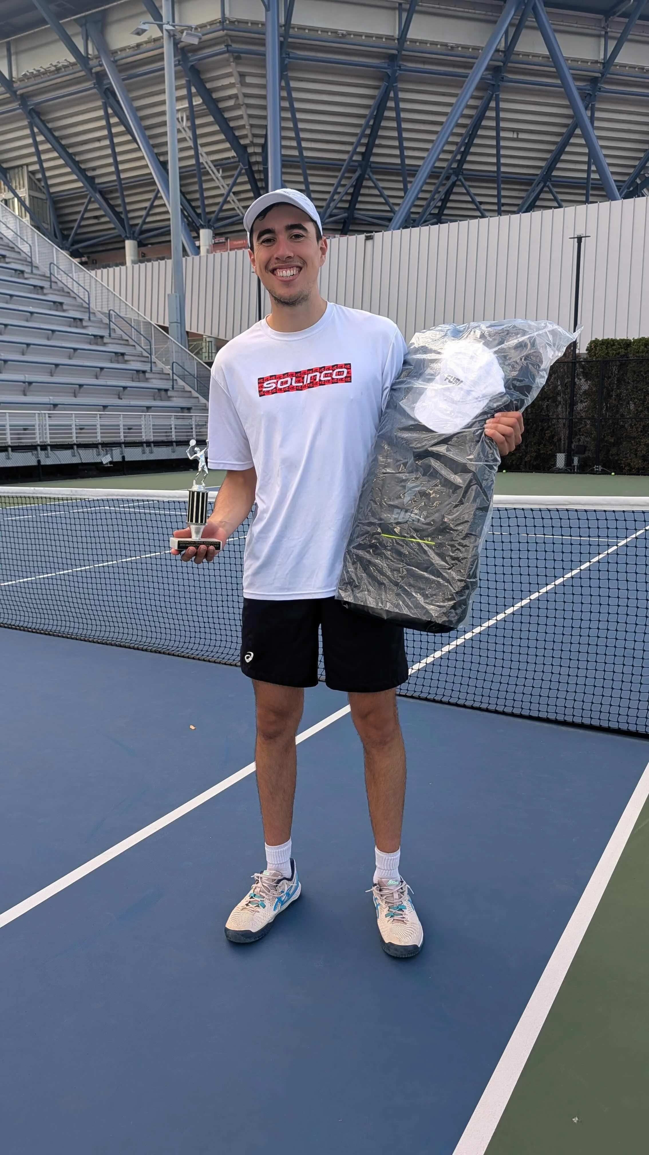 A young man standing on a tennis court holding a trophy in one hand and a large bag of prizes in the other. He is smiling and wearing a white tennis cap, a white Solinco shirt, black shorts, and tennis shoes.