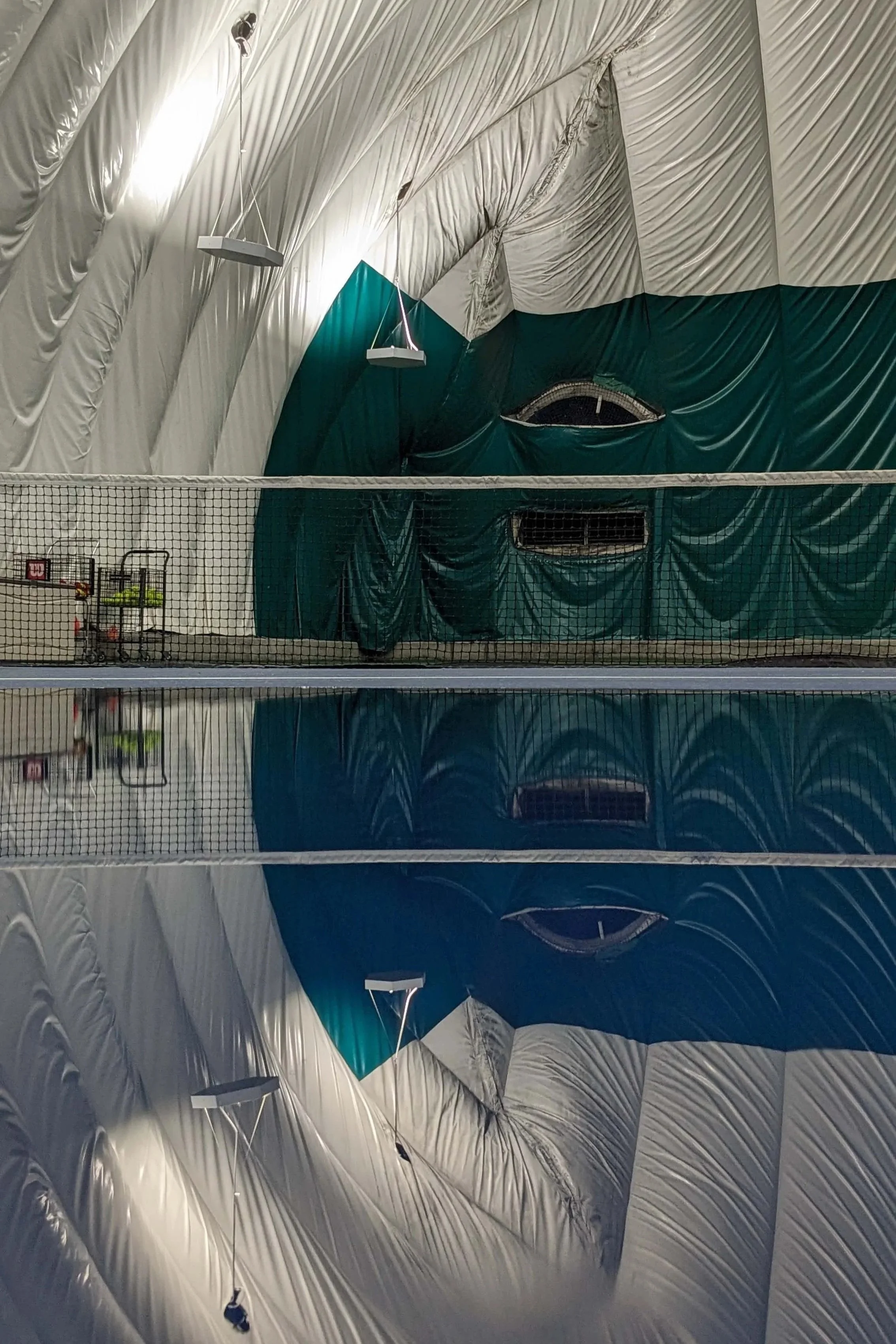 Indoor tennis court with a green curtain wall, net in the middle, and reflective polished floor showing the curtain and ceiling.