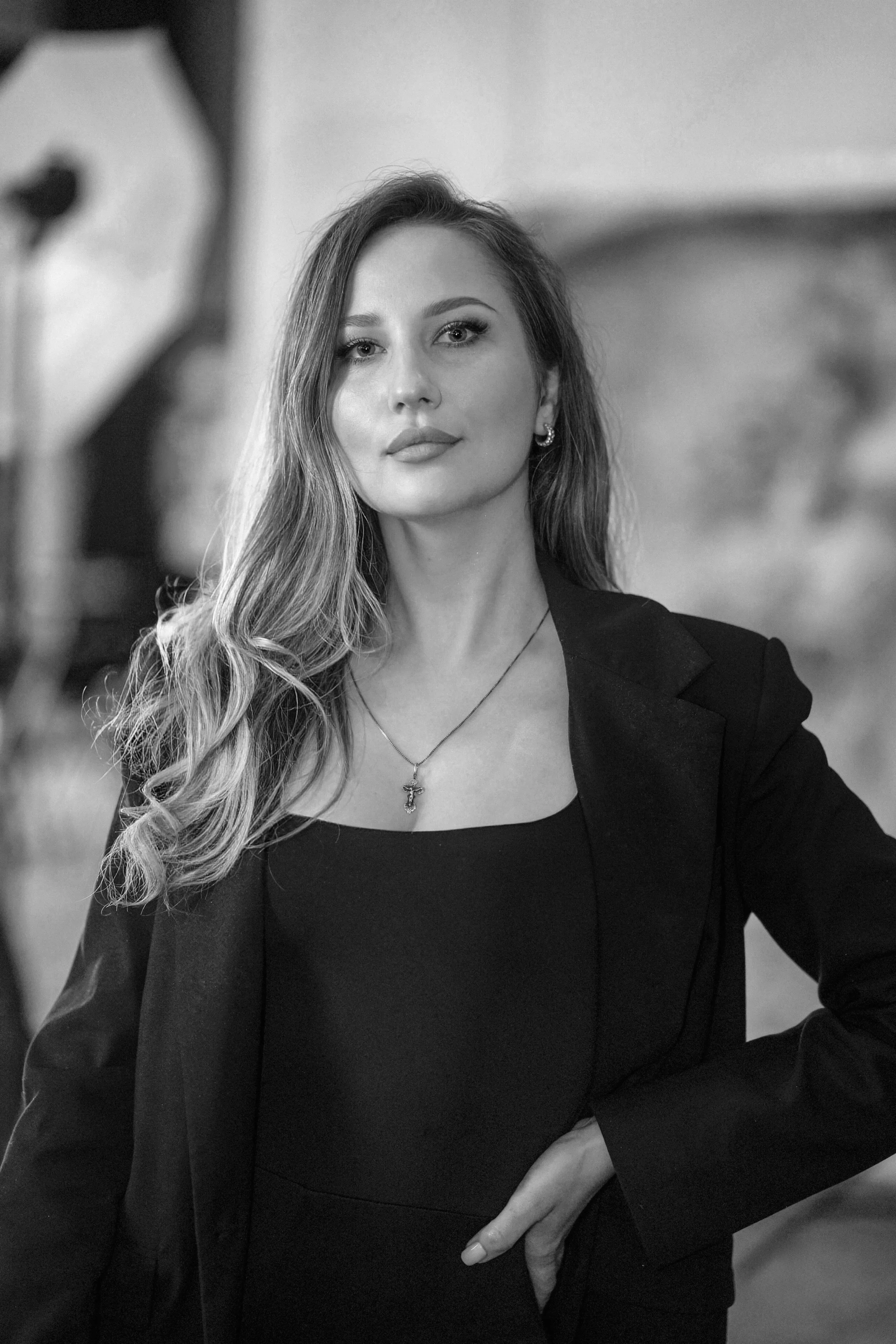 A woman in a black blazer and dress, with long wavy hair, wearing earrings and a necklace, standing indoors with a blurred background.