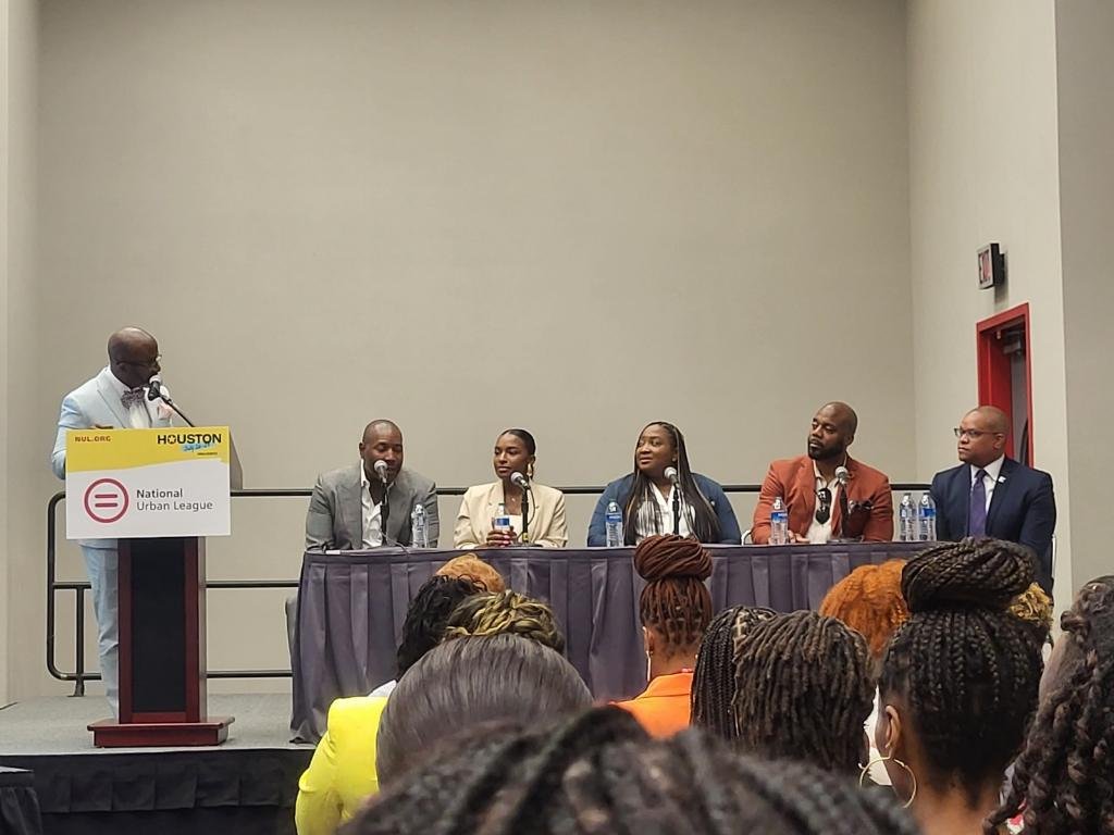 Panel of five individuals seated at a table on stage, with one standing at a podium speaking into a microphone, during an event organized by the National Urban League in Houston.