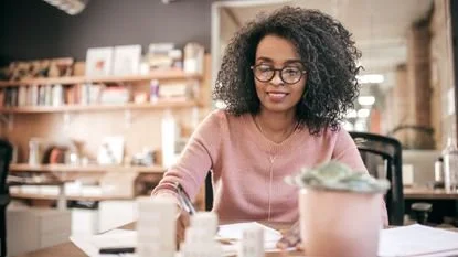 Woman with curly hair and glasses smiling at a desk in a cozy, well-lit cafe or workspace, with books and shelves in the background.