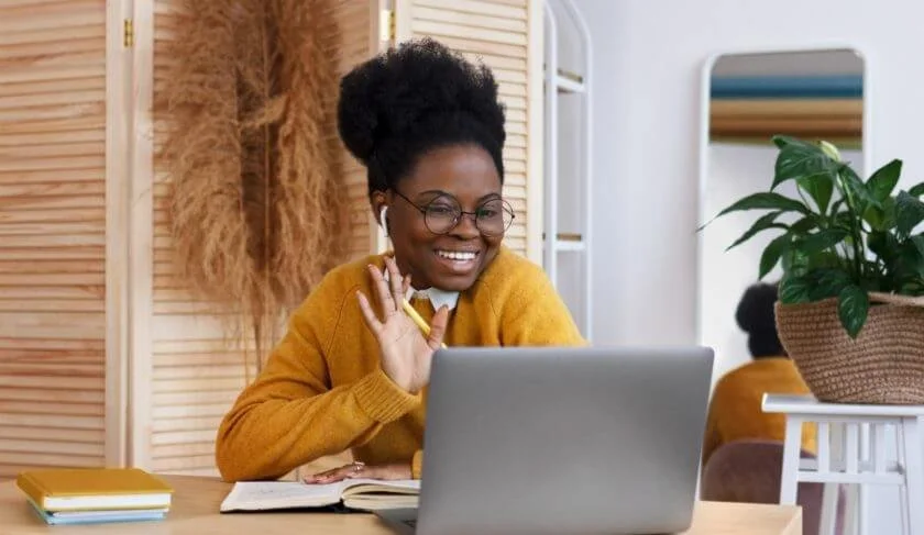 A woman with glasses and a natural hairstyle smiling while waving at a laptop on her desk, surrounded by notebooks and a potted plant.