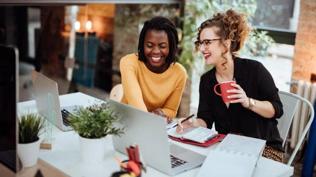 Two women sitting at a table with laptops, having a conversation and smiling, in an office or cafe setting.