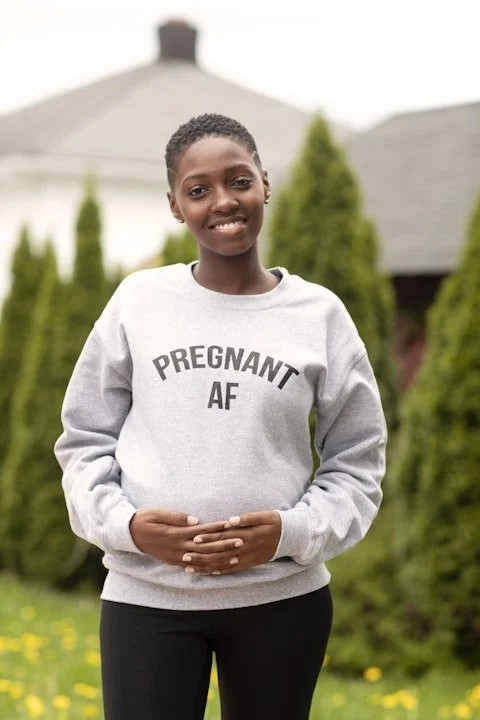 Young woman standing outdoors in front of greenery, smiling, wearing a gray sweatshirt with the words 'PREGNANT AF' printed on it.