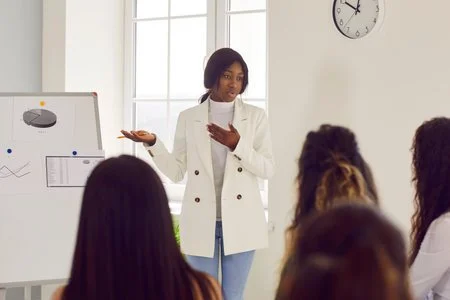 Woman giving a presentation to a group of people in a bright room with a whiteboard and a clock on the wall.
