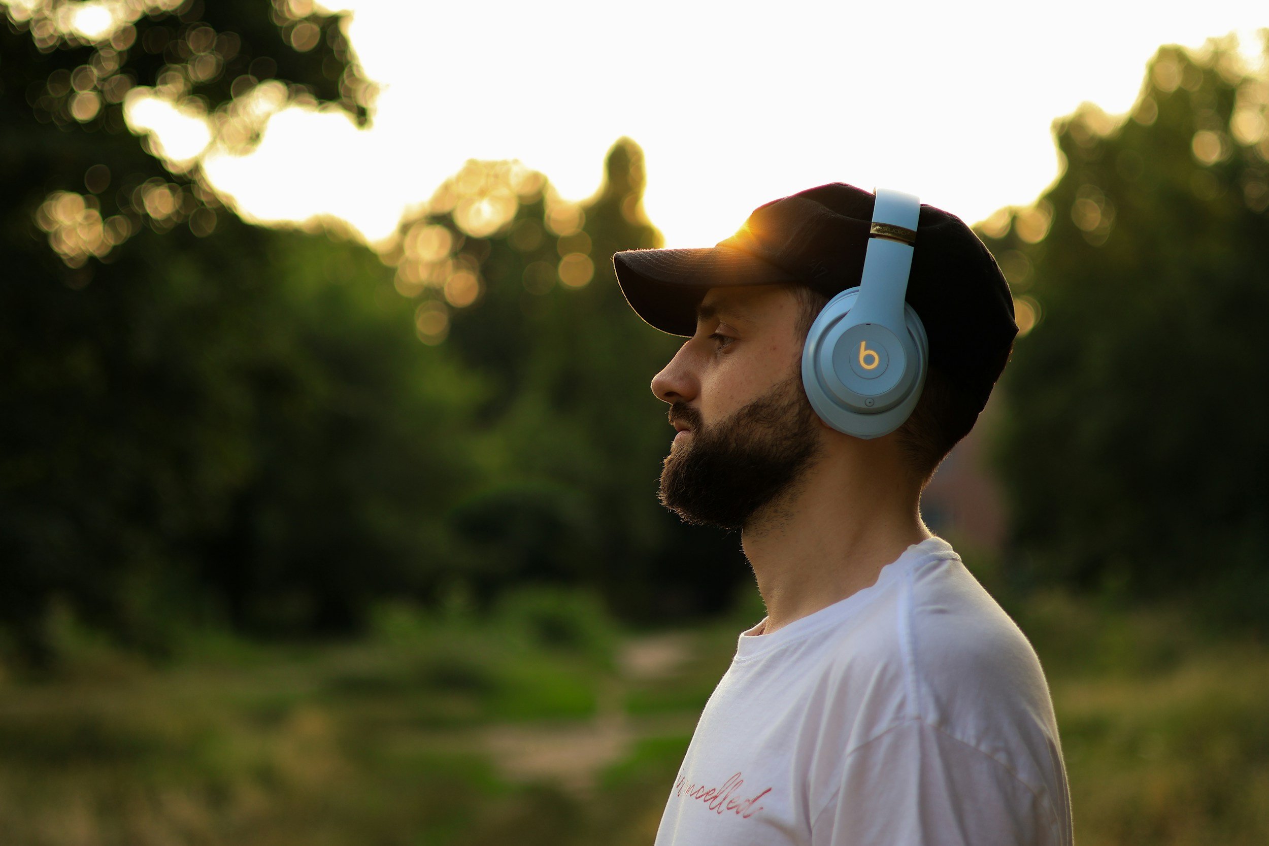 Man with a beard, wearing a black baseball cap and white t-shirt, listening to blue Beats headphones outdoors during sunset.