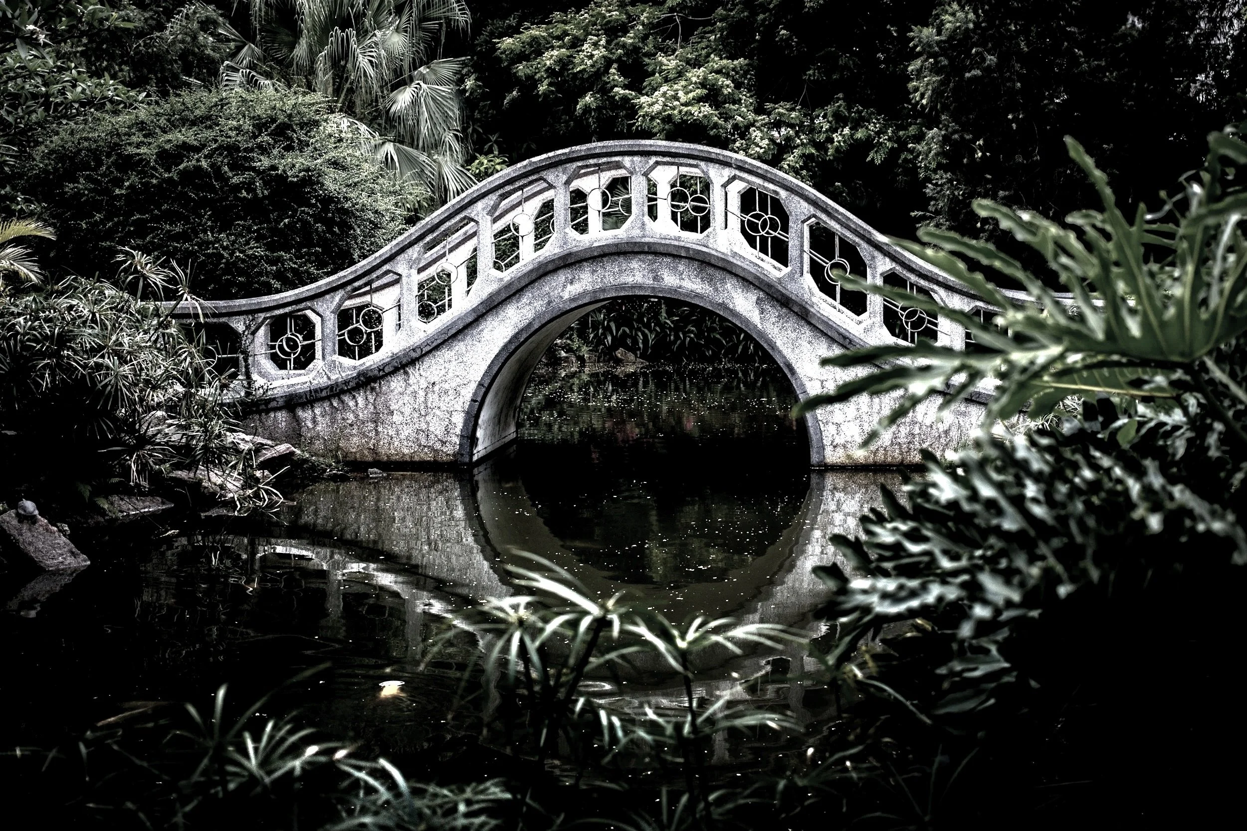A stone bridge with geometric patterns spans over a small body of water, surrounded by lush foliage and trees, with some leaves in the foreground and dense greenery in the background.