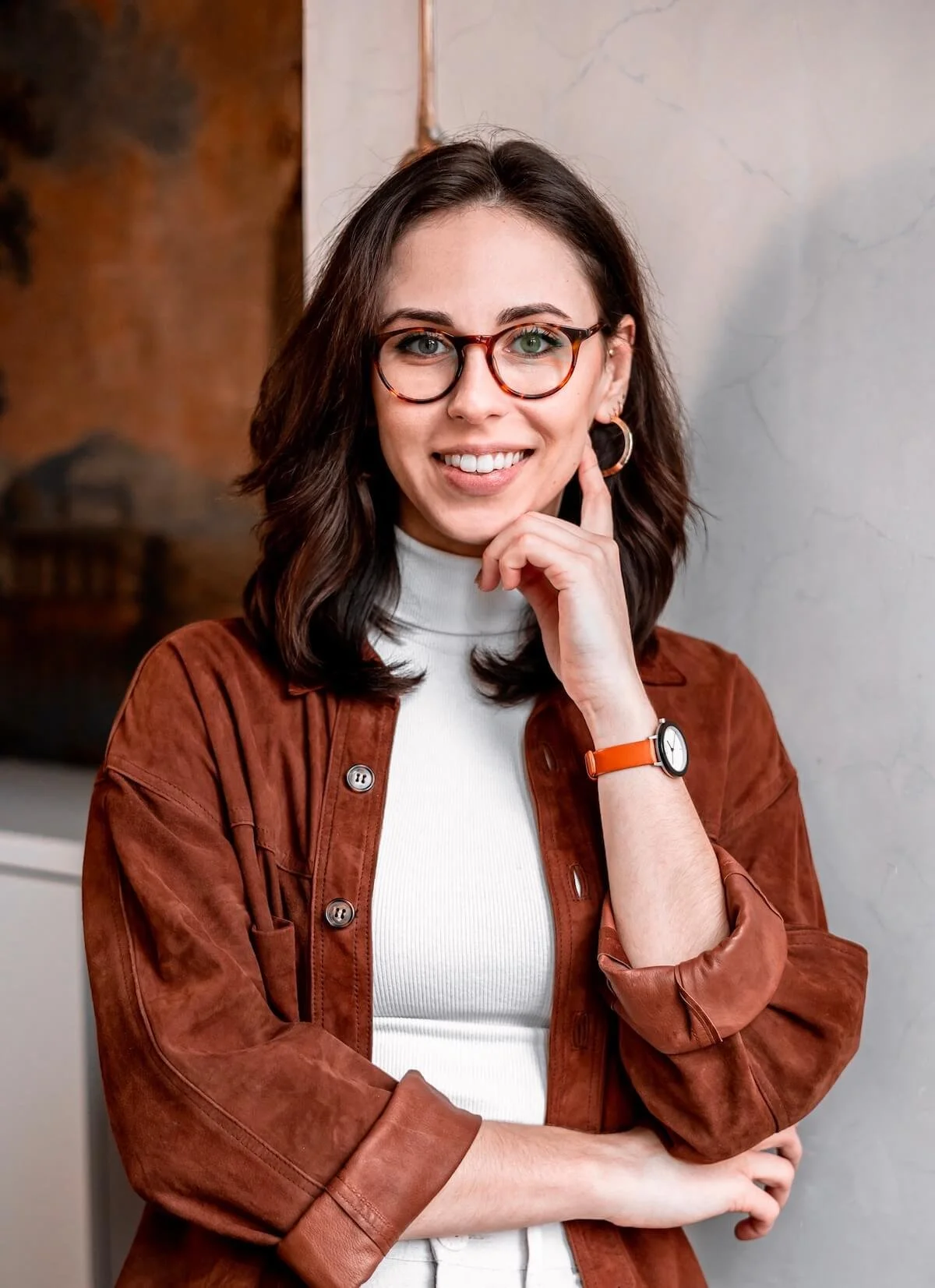 Eine junge Frau mit dunklen, gewellten Haaren, Brille und Ohrringen, trägt ein weißes Rollkragenshirt unter einem braunen Jacke und trägt eine Armbanduhr, steht vor einer Wand.