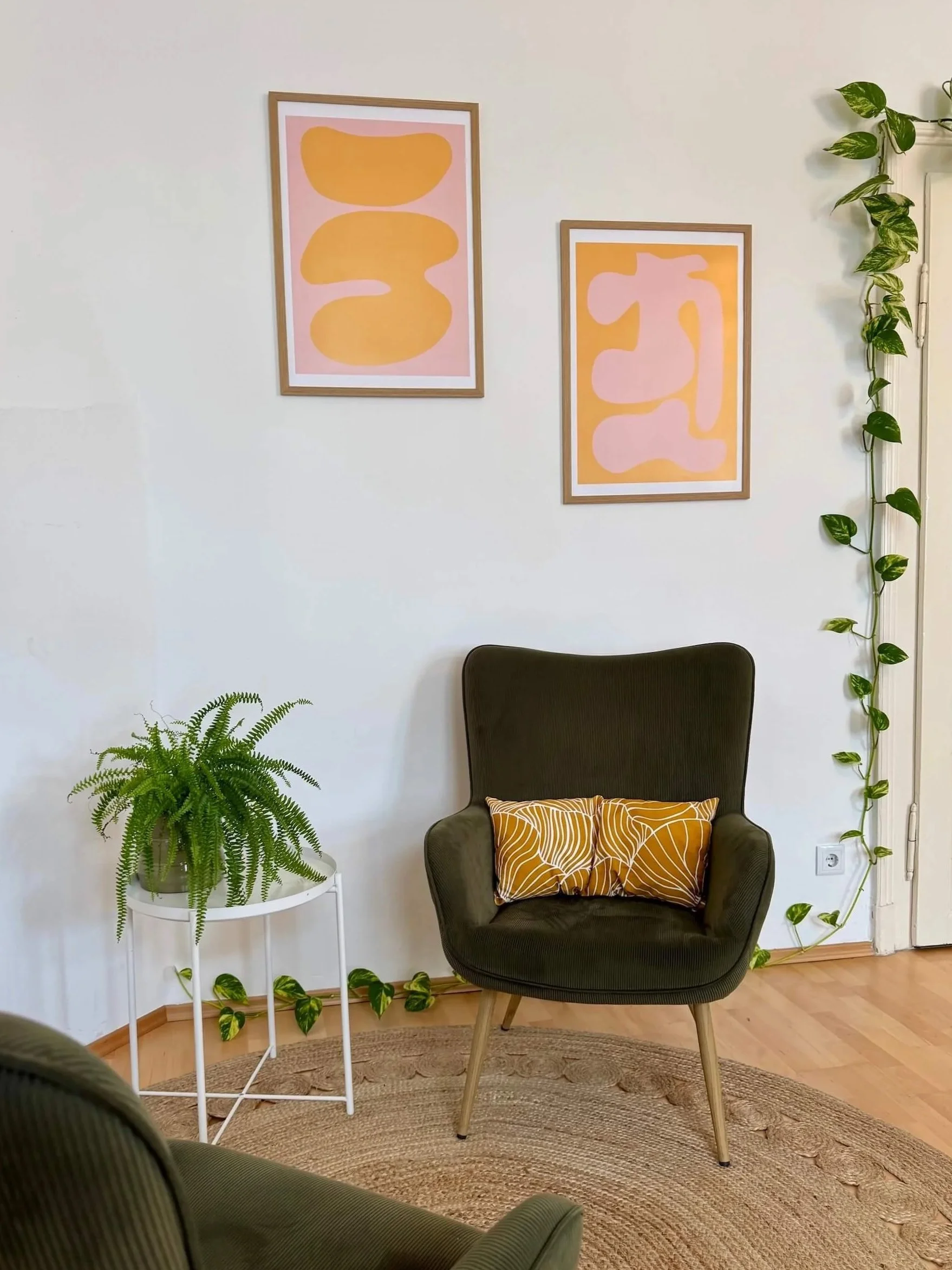 Ein gemütliches Zimmer grünen Sessel, einem kleinen weißen Beistelltisch mit einer Topfpflanze, Wanddekoration mit abstrakten Kunstwerken in Pastellfarben, und vielen grünen Pflanzenranken an der Wand.