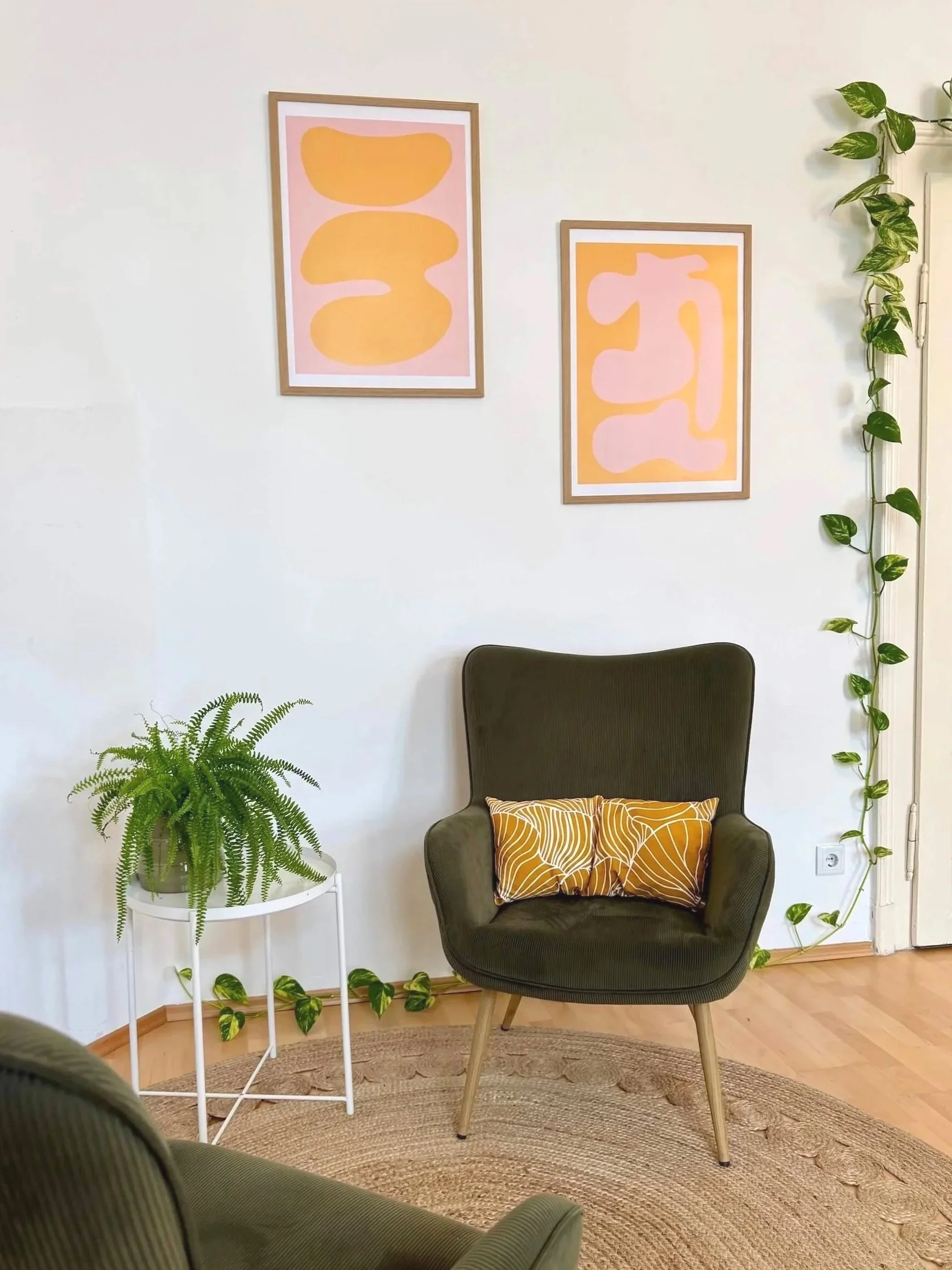 Ein gemütliches Zimmer mit einem grünen Sessel, einem kleinen weißen Beistelltisch mit einer Topfpflanze, Wandkunst mit abstrakten Formen in Pastellfarben, Pflanzenranken an der Wand und auf dem Boden.