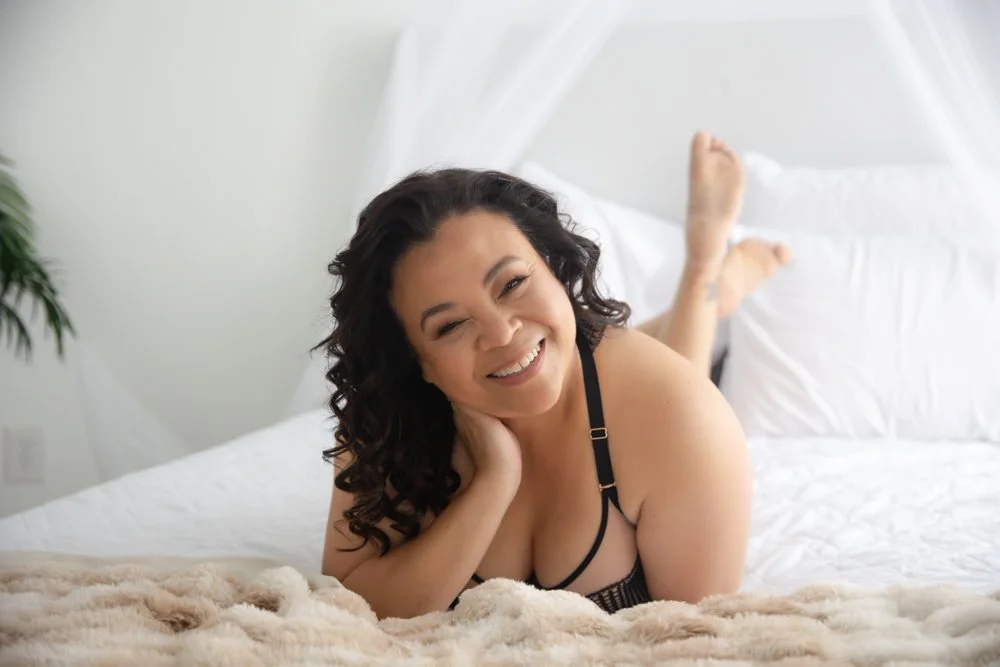 Woman with dark curly hair in black lingerie smiles while reclining on a cream blanket in a bright, minimalist bedroom with soft natural light.