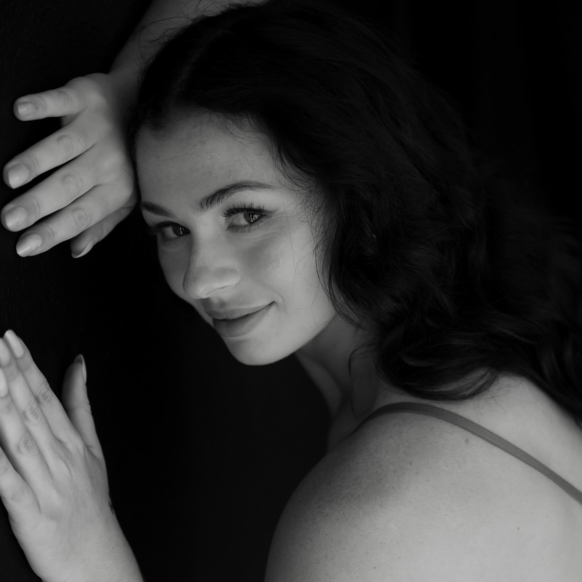 Professional portrait of a woman with dark hair posing with hands near face against a dark background, showcasing studio portrait photography and artistic lighting.