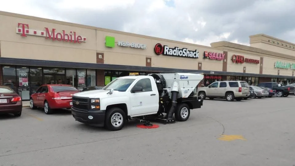 A white street cleaning truck parked in a shopping center parking lot in front of a strip mall with stores including RadioShack, Sally, and T-Mobile.