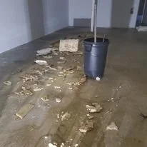 A large amount of water and debris, including broken tiles and mud, spilled across the floor in what appears to be a damaged or flooded indoor area, with a trash bin nearby.