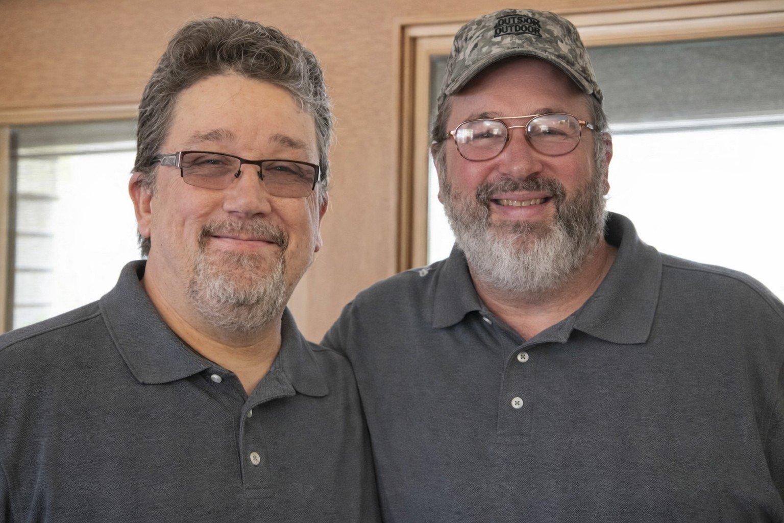 Two middle-aged men with glasses and beards smiling and posing together, wearing dark polos, standing indoors in front of a window with wooden framing.