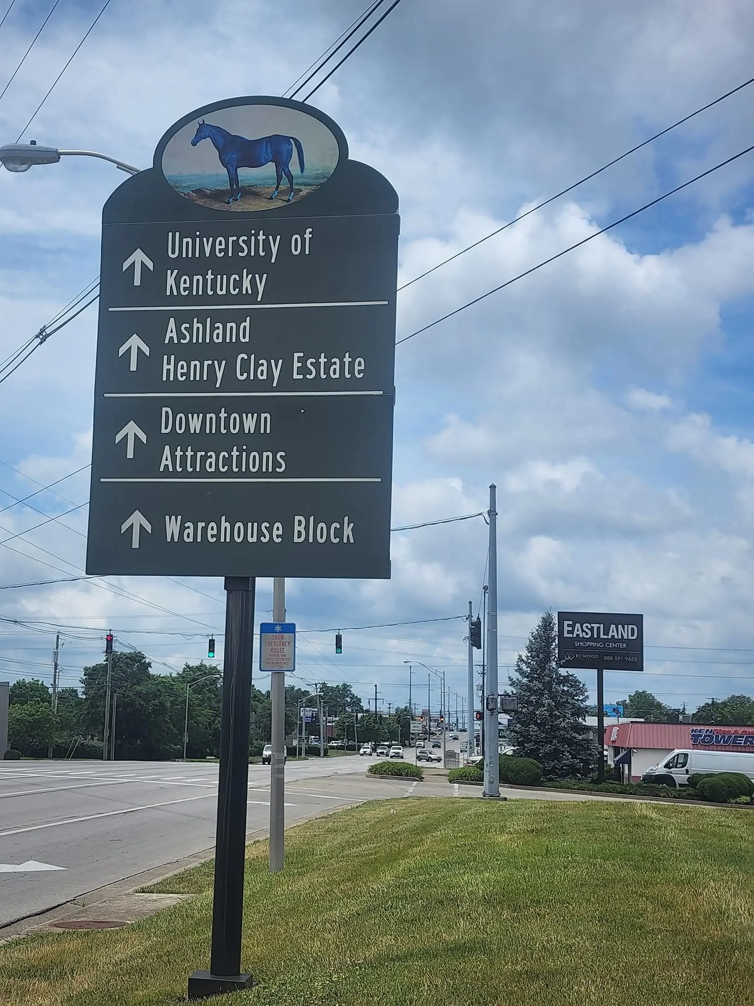 Street sign with a blue horse logo at the top, directing to University of Kentucky, Ashland Henry Clay Estate, Downtown Attractions, and Warehouse Block, with a shopping center sign reading Eastland in the background.