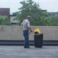 Man standing next to a black trash can on a rooftop, holding a cleaning tool with a yellow cloth.