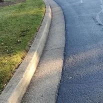 A curb and sidewalk next to a grassy area and a wet road.