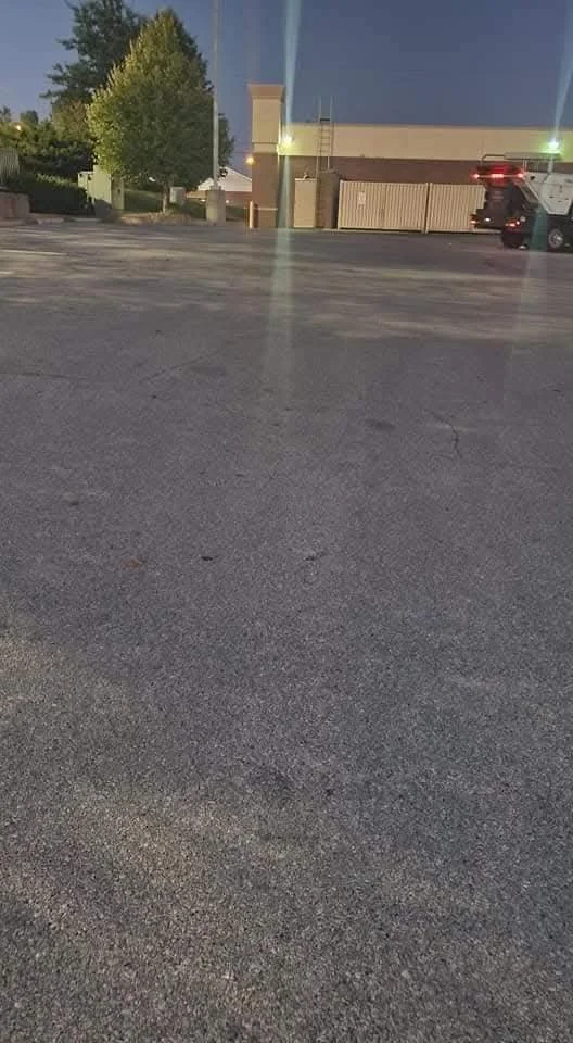 Empty parking lot at dusk with a building, trees, and a few parked cars in the background.