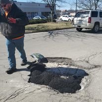 A person standing next to a large hole in the pavement filled with hot asphalt, in a parking lot.