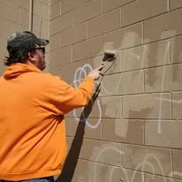 A person wearing an orange hoodie and camouflage hat removing graffiti on a beige brick wall.